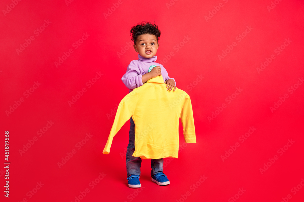 Full length body size view of attractive funny kid holding yellow knitted swetaer store isolated over bright red color background Full length body size view of attractive funny kid holding yellow knitted swetaer store isolated over bright red color background