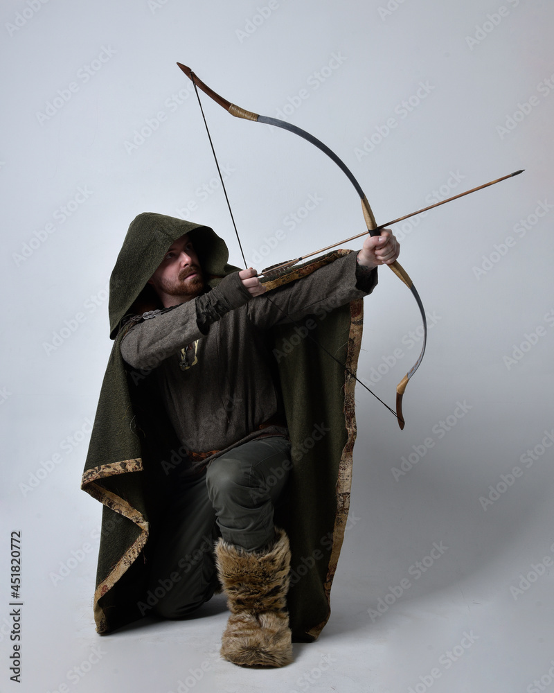 Full length  portrait of  young handsome man  wearing  medieval Celtic adventurer costume with hooded cloak, holding a archery bow and arrow, isolated on studio background.