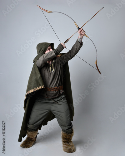 Full length portrait of young handsome man wearing medieval Celtic adventurer costume with hooded cloak, holding a archery bow and arrow, isolated on studio background.