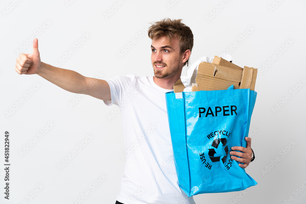 Young blonde man holding a recycling bag full of paper to recycle isolated on white background giving a thumbs up gesture Young blonde man holding a recycling bag full of paper to recycle isolated on white background giving a thumbs up gesture