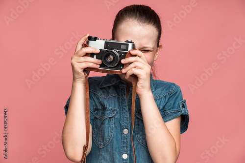 Keep smile. Little girl holding vintage photo camera and shooting somebody on it. Girl satisfied with photo shoot. Indoor studio shot isolated on pink background