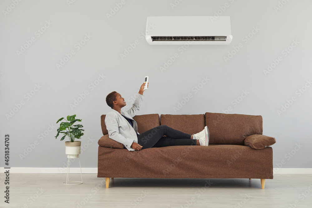 Happy young black woman who's lounging on comfortable brown sofa in  minimalist home interior looks above at air conditioner and presses power  switch on remote control. Modern AC system concept Stock Photo