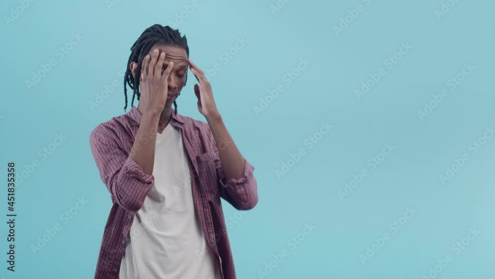 Tense black man with dreadlocks rubs hand on chin and sighs wearily standing on a blue studio background. Concept of confusion and anxiety. Tense black man with dreadlocks rubs hand on chin and sighs wearily standing on a blue studio background. Concept of confusion and anxiety.