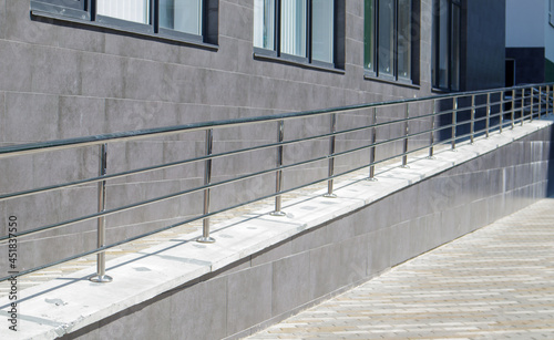 A ramp with stainless steel handrails for wheelchairs, bicycles and strollers with children in front of the building. Handrail for lifting a wheelchair. Shiny stainless steel handrail.
