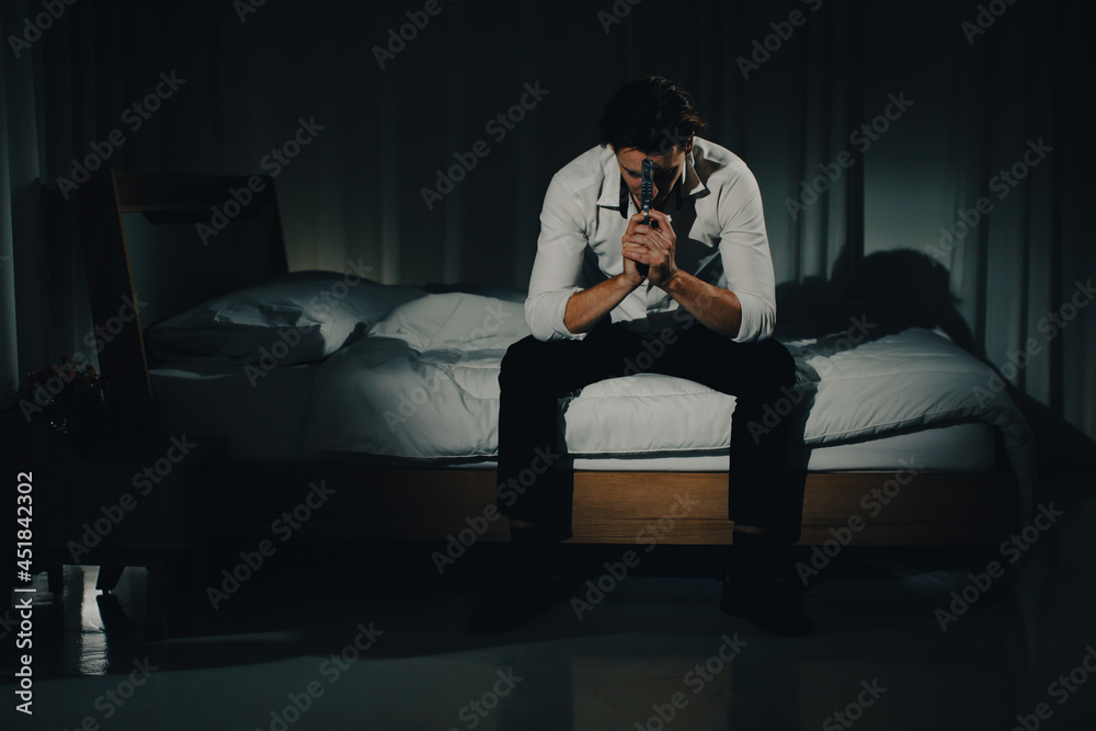 A young man in casual business clothes sitting alone in bedroom holding a pistol in a thoughtful and serious mind look like going to kill himself. Concept of hopelessness and give up people.