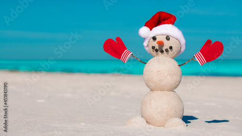 Snowman on the beach. Sandy Christmas snowman in red Santa hat and mittens or gloves. Smiley Snowman at sunny beach. Holiday concept for Happy New Year post cards. Florida Winter. Ocean or Sea water.