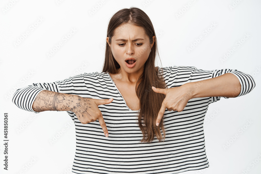 Portrait of woman pointing fingers down with shocked face. Female model with tattoos and piercing looking overwhelmed and frustrated, white background Portrait of woman pointing fingers down with shocked face. Female model with tattoos and piercing looking overwhelmed and frustrated, white background