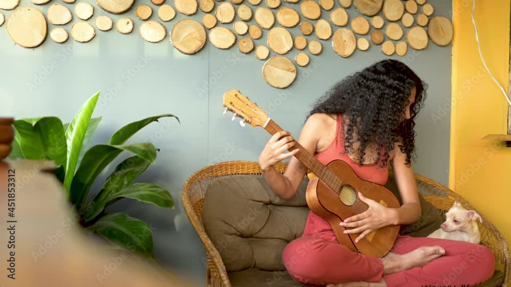 Mujer morena de cabello rizado tocando guitarra con su mascota en una banca en un hostal