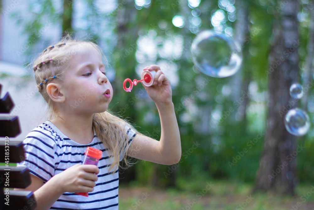 A little blonde girl in a striped T-shirt on vacation sits on a park bench in good weather and happily blows soap rainbow bubbles close-up. There are buildings and green trees all around.