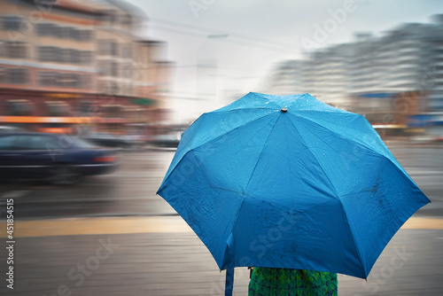 Woman with wet umbrella walking in rainy day on city street. Woman under umbrella  waiting to cross the road on pedestrian crossing. Surface of blue umbrella with rain drops