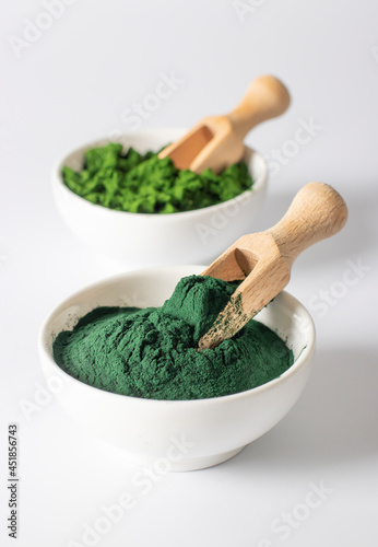 Organic algae chlorella and spirulina in powder form in white bowls with wooden scoops on a white background.