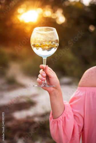 A girl in a pink dress holds a beautiful glass of water and flowers in the setting sun. Glare, summer, nature. Content