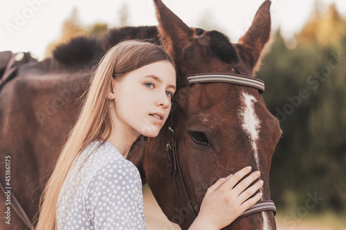 A beautiful young girl with long hair stands next to a horse in nature in the summer © Olya Komarova