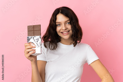 Teenager Ukrainian girl isolated on pink background taking a chocolate tablet and happy