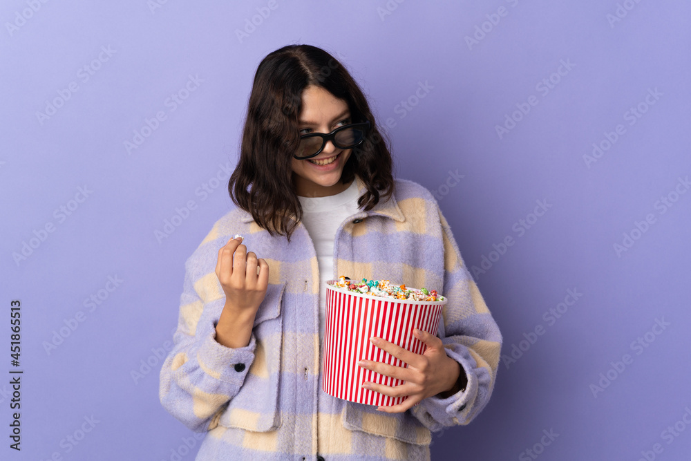 Teenager Ukrainian girl isolated on purple background with 3d glasses and holding a big bucket of popcorns while looking side