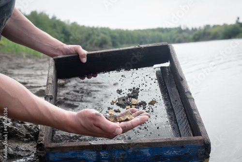 Treasure hunter is picking a golden ore from dirty metal grid on the river water background. Sifting gold concept. photo