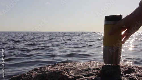 Close-up, dark silhouette of a travel thermos mug of tea or coffee, on a stone, against background of sunrise or sunset, in the rays of sun light and glimming water waves
