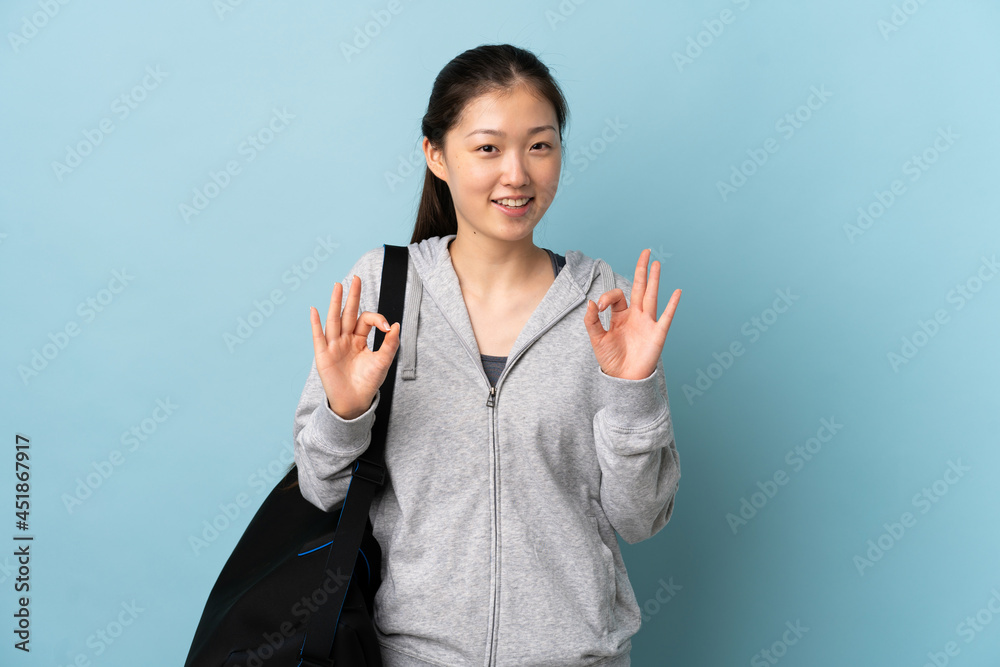 Young sport Chinese woman with sport bag over isolated blue background showing ok sign with two hands Young sport Chinese woman with sport bag over isolated blue background showing ok sign with two hands