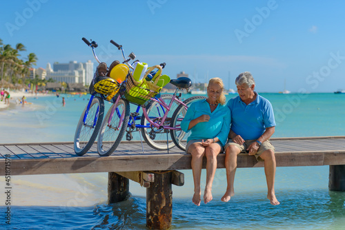 Happy senior couple exercising with bicycles on the beach on a sunny day, sitting on a pier © TRAVEL EASY