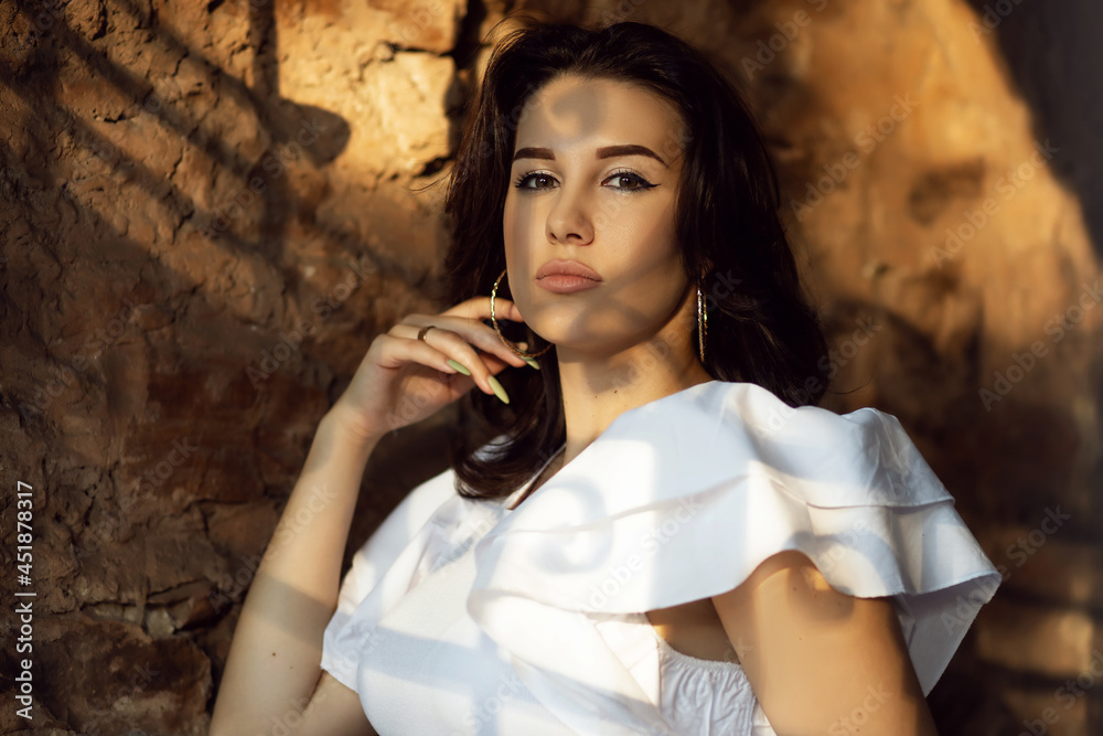 Obraz premium Lovely young brunette woman posing against the background of an old wall with shadows on her face during sunset. Stylish portrait of a girl in a white T-shirt. A woman touches her face