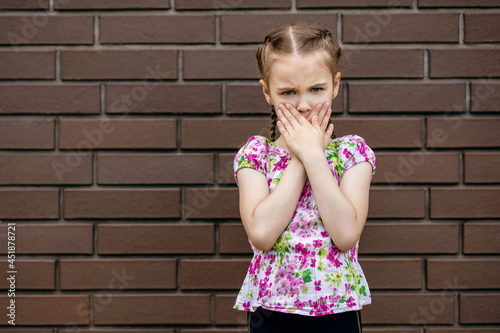 The little girl covers her mouth with her palms against the background of a brick wall. the pigtailed child frowned and displeased. Portrait of an offended emotional child. Free space for text photo