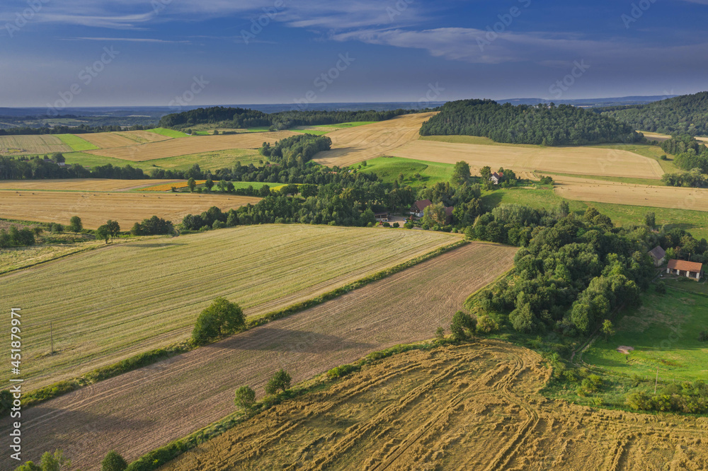 Sudeten foothills. Undulating terrain covered with arable fields, meadows, clumps of trees. In the distance you can see the buildings of the village and mountains on the horizon. Photo from the drone.