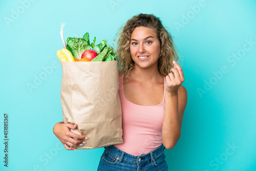 Girl with curly hair holding a grocery shopping bag isolated on green background making money gesture