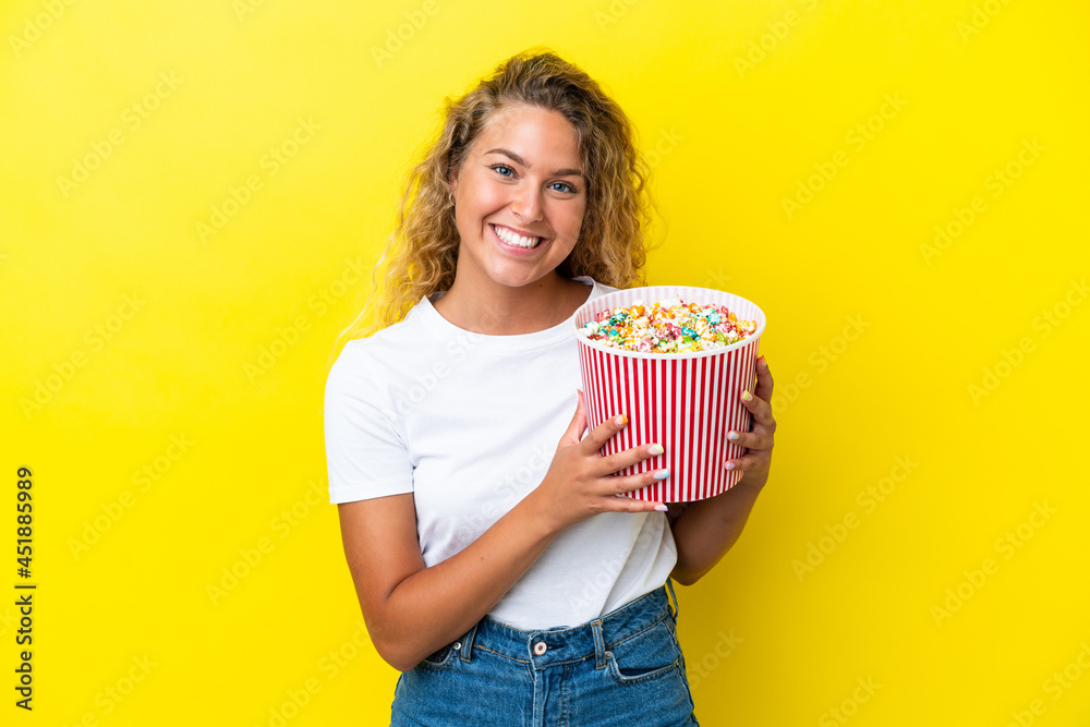 Girl with curly hair isolated on yellow background holding a big bucket of popcorns