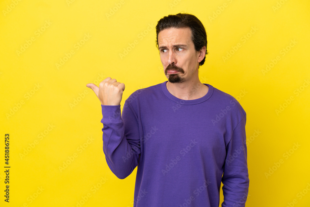 Young caucasian man isolated on yellow background unhappy and pointing to the side Young caucasian man isolated on yellow background unhappy and pointing to the side