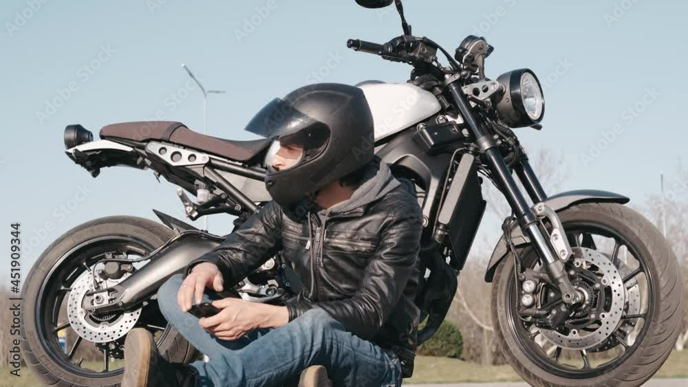 Young attractive motorcyclist with black helmet and black leather jacket on the street using smartphone Young attractive motorcyclist with black helmet and black leather jacket on the street using smartphone