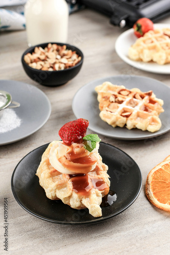 Delicious Homemade Croissant Waffle with Cream, Maple Syrup, Chopped Almond, and Strawberry. Served on Black Plate, Brown Background.
