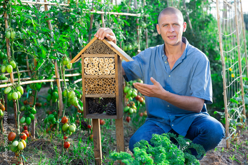 Portrait of positive man next to an insect hotel in form of wooden birdhouse in the garden. Concept handmade © JackF