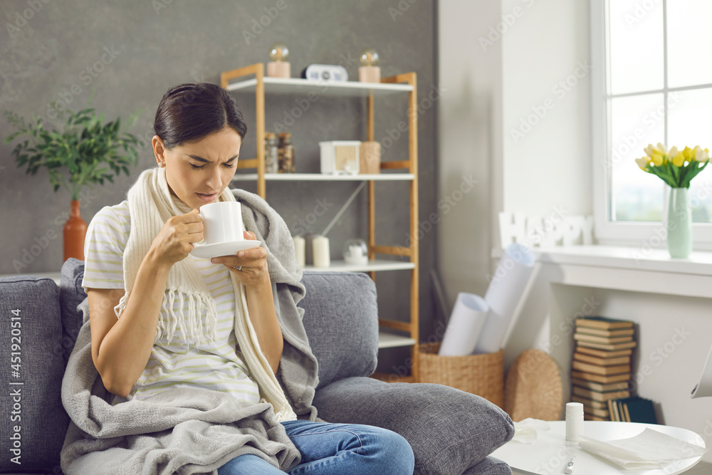 Sad unhappy sick woman feeling unwell and suffering from common cold or seasonal flu with fever holding mug and drinking warm tea while sitting on couch at home wrapped in scarf and plaid blanket Sad unhappy sick woman feeling unwell and suffering from common cold or seasonal flu with fever holding mug and drinking warm tea while sitting on couch at home wrapped in scarf and plaid blanket