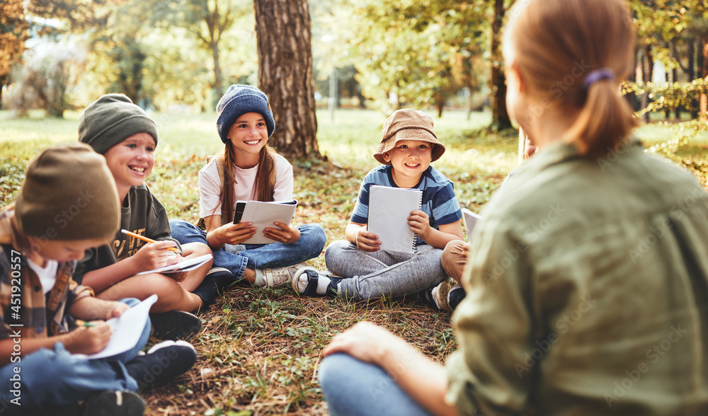 Smiling children and female teacher sitting in circle on grass in forest and talking about nature Smiling children and female teacher sitting in circle on grass in forest and talking about nature