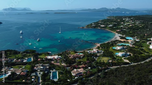 Touristic sailing ship boats in a beautiful Mediterranean hotel beach seaside bay with crystal clear blue turquoise ocean water in Sardinia, Italy and little waves. Holiday vacation aerial drone above