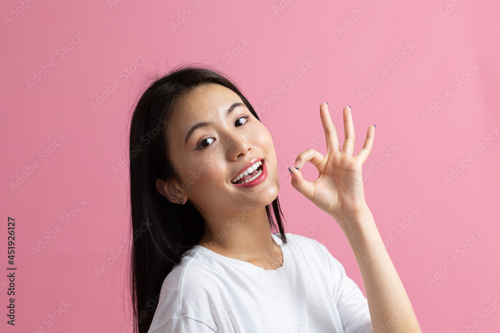 Smiling asian girl showing Ok gesture. Beautiful young woman with dark hair wearing white t-shirt. Female looking at camera isolated on pink background. Studio shoot Smiling asian girl showing Ok gesture. Beautiful young woman with dark hair wearing white t-shirt. Female looking at camera isolated on pink background. Studio shoot