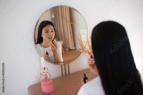 Beautiful Asian woman front of mirror applying make up lips with pink lipstick at living room.