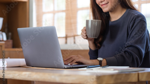 A young woman freelancer drinking coffee while using laptop computer for working online at home