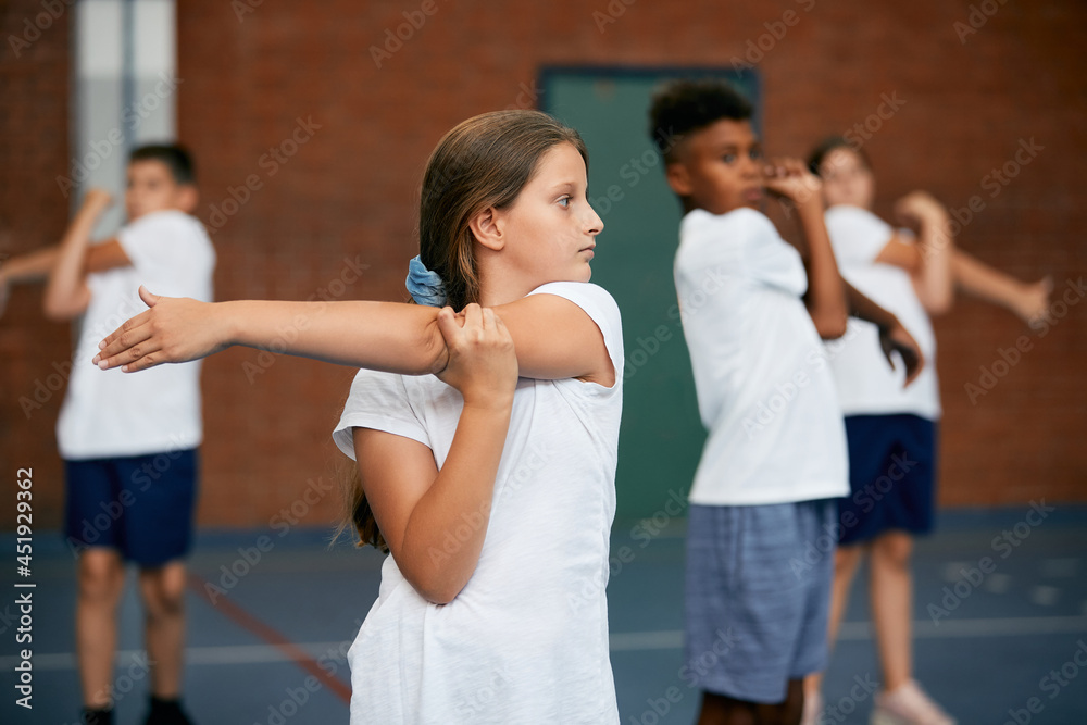 Little girl stretches her arm while having physical education class with her friends at school gym.