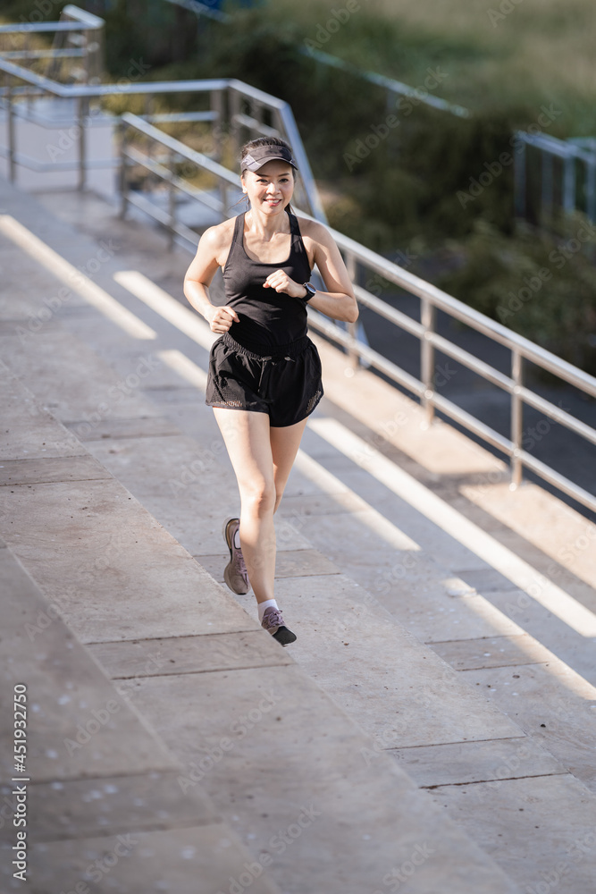 A healthy happy Asian woman runner in black sport outfits jogging in the natural city park under evening sunset