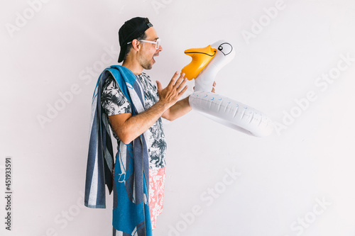 Young man in a floral shirt, cap, towel and glasses in summer, with a float duck making joyful gestures, in daylight on a white wall photo