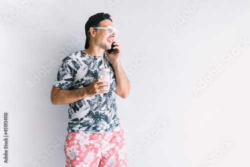 Young man in a floral shirt, cap and glasses in summer talking on his smart phone and drinking a cocktail with a straw, in daylight on a white wall photo