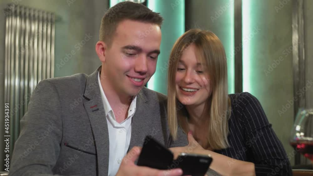 An attractive couple is sitting at a table using a smartphone, browsing the web, discussing online purchases, ordering food from a restaurant for a romantic dinner An attractive couple is sitting at a table using a smartphone, browsing the web, discussing online purchases, ordering food from a restaurant for a romantic dinner
