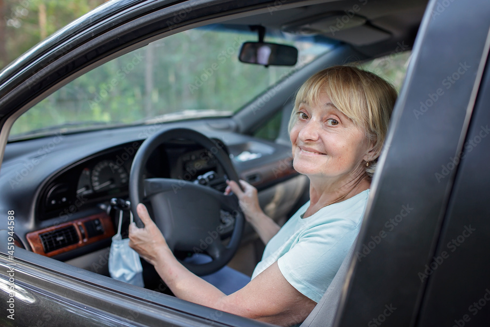Happy senior woman driving a car, medical mask hanging on the steering wheel, solo traveler, independent and active retirement