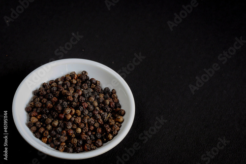 Black Pepper isolated in a white bowl on a black background is used as an ingredient in Indian food. 