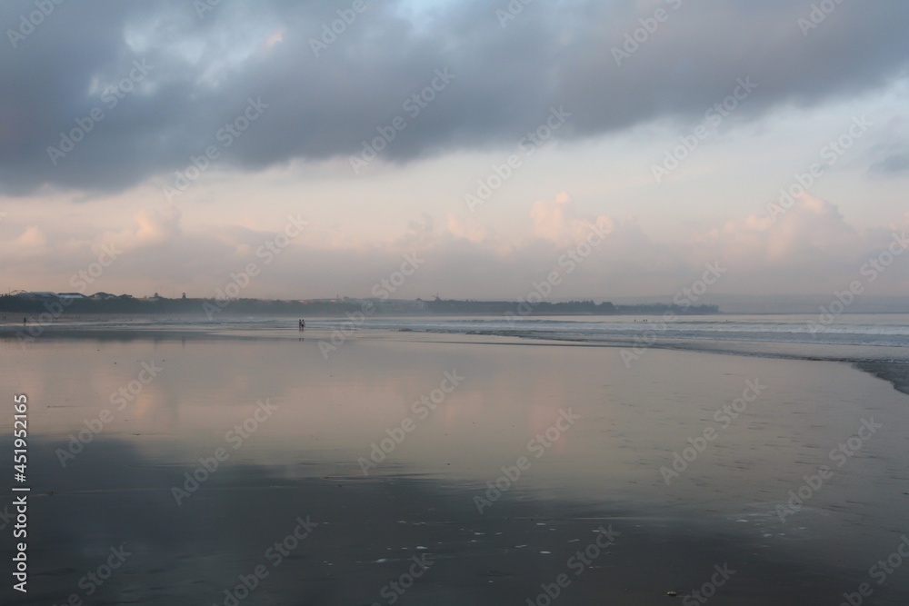 Scenic view of beach in morning at sunrise with mirror reflection on wet sand. Clouds variation in blue sky and people walking on beach. Panorama view.