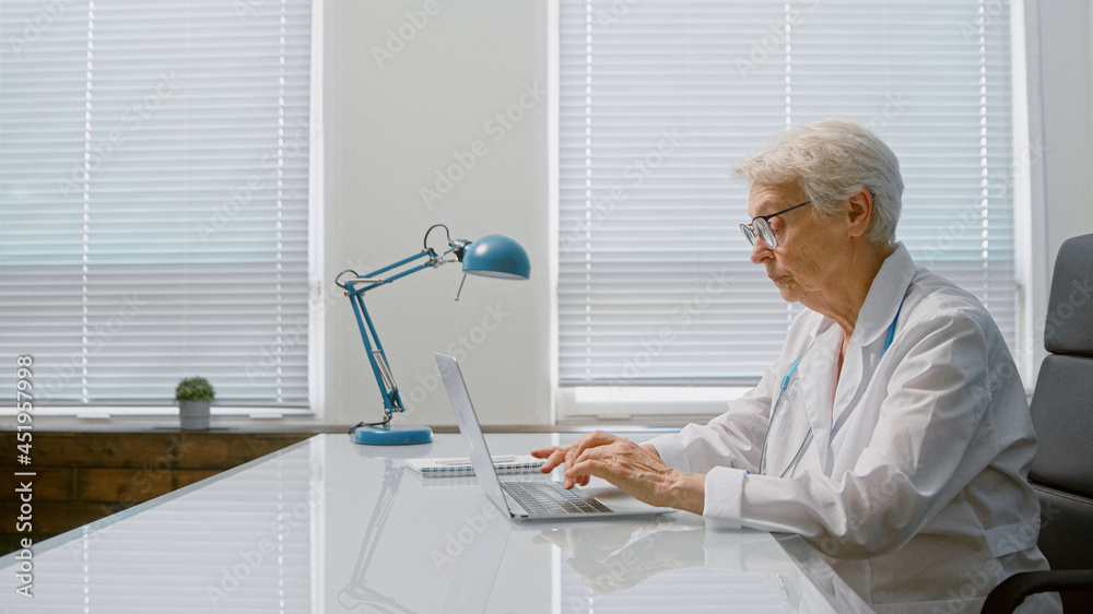 Senior short grey haired lady doctor writes letter on contemporary laptop sitting at table in spacious clinic