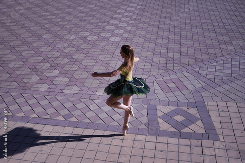 Adult female Hispanic classical ballet dancer in green and black tutu with coins, dancing in the middle of a plaza casting her shadow on the ground.