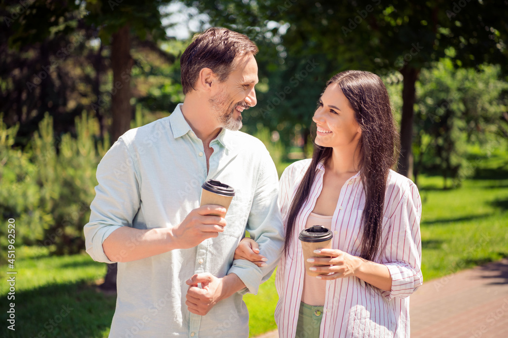 Photo of sweet pretty mother father dressed casual outfit holding arms walking holding beverages smiling outside city park Photo of sweet pretty mother father dressed casual outfit holding arms walking holding beverages smiling outside city park