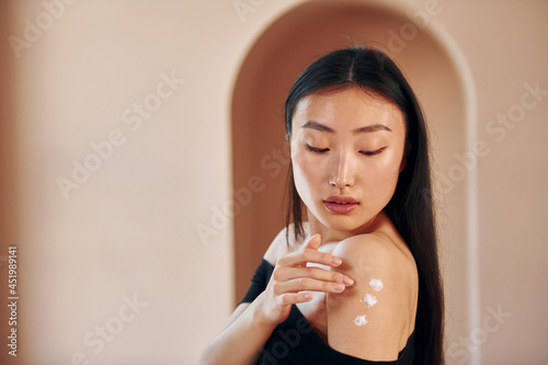 Fototapeta Naklejka Na Ścianę i Meble -  Uses beauty cream. Young serious asian woman standing indoors